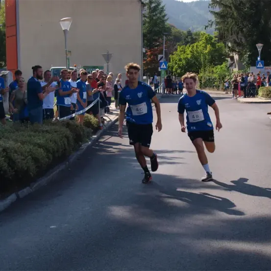 Zwei Läufer mit den Nummern 4C und 4B laufen auf einer Straße, hinter ihnen steht eine Menge von Zuschauern. Die Menge jubelt und hält eine Barriere. Bäume und Gebäude befinden sich an den Seiten der Straße.