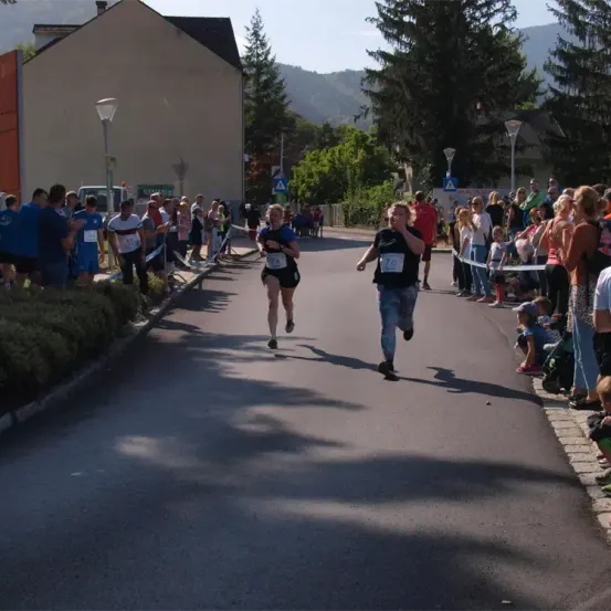Zwei Läufer nehmen an einem Rennen teil, das von einer Menge beobachtet wird. Bäume und Gebäude sind im Hintergrund zu sehen.