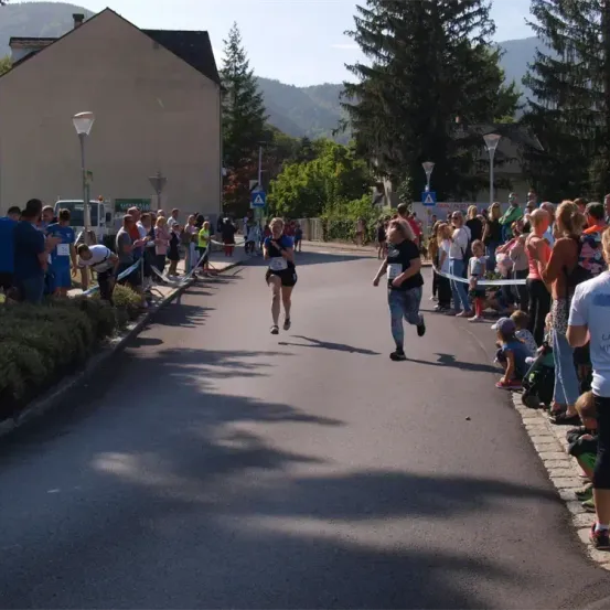 Zwei Frauen laufen in einem Marathon, umgeben von einer Menge von Zuschauern, Gebäuden und Bäumen.