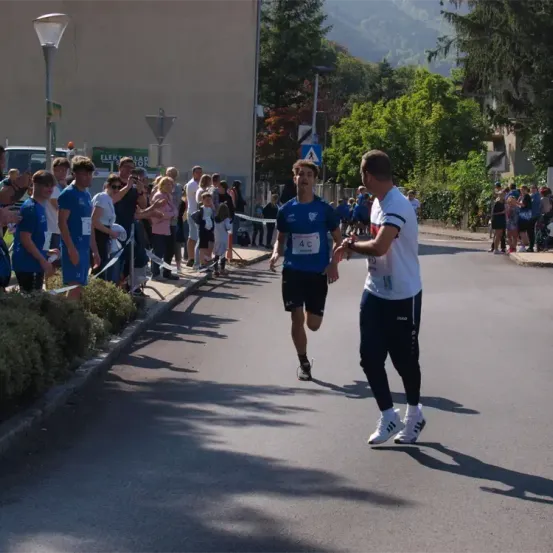 Ein Marathonläufer mit einer blauen Nummer 4 auf dem Trikot läuft auf der Straße, ein Mann in Weiß nähert sich ihm, um ihm etwas zu übergeben.