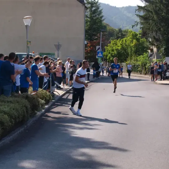 Ein Läufer sprintet die Straße hinunter, während Zuschauer vom Bürgersteig zujubeln. Hohe Bäume und Gebäude säumen die Straße.