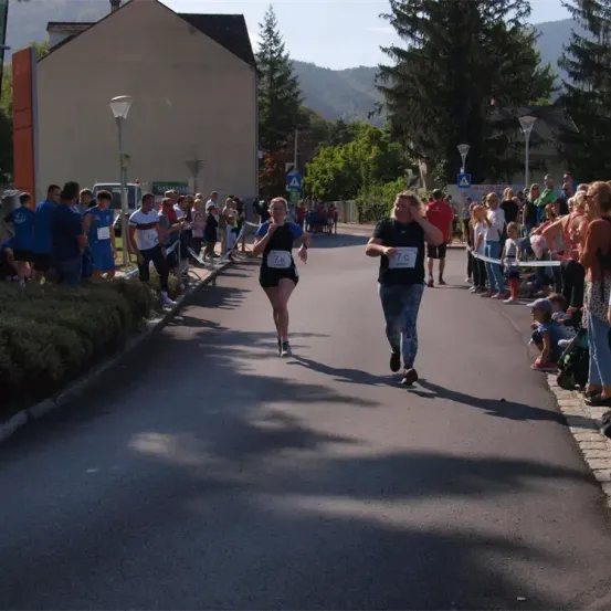 Ein Marathon findet auf einer Straße statt. Zwei Läufer, einer mit der Nummer 7B und der andere mit TC, laufen vor einer Menge. Zuschauer stehen am Straßenrand, und Straßenlaternen sind sichtbar.
