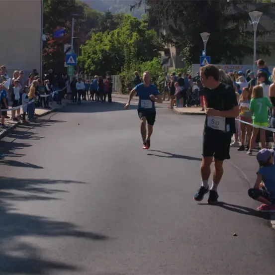 Ein Läufer in einem blauen Shirt mit einer Nummernplakette läuft mitten auf einer Straße, während Zuschauer an der Seite stehen. Ein Mann in Schwarz steht in der Nähe und beobachtet.