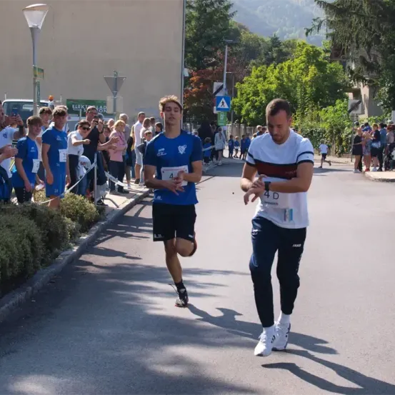 Zwei Männer laufen in einem Rennen, einer in einem blauen Shirt und der andere in einem weißen Shirt. Zuschauer beobachten von der Straßenseite aus.