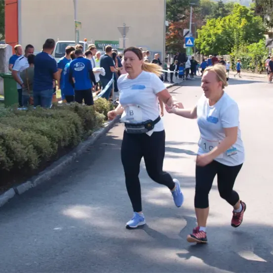 Zwei Frauen laufen in einem Marathon, eine mit der Nummer B und die andere mit der Nummer 10. Sie halten Händchen und lächeln. Dahinter stehen Leute und beobachten.
