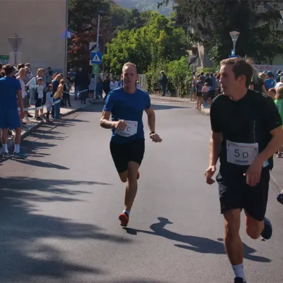Zwei Männer laufen in einem Marathon, einer mit der Nummer 5D und der andere mit C, Zuschauer säumen die Straße.
