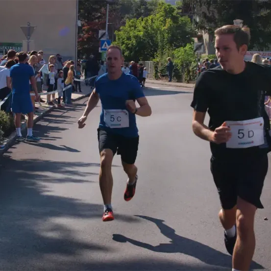 Zwei Männer laufen in einem Rennen, einer mit der Nummer 5C, der andere mit 5D. Zuschauer säumen die Straße hinter ihnen, und Bäume und Gebäude sind im Hintergrund.