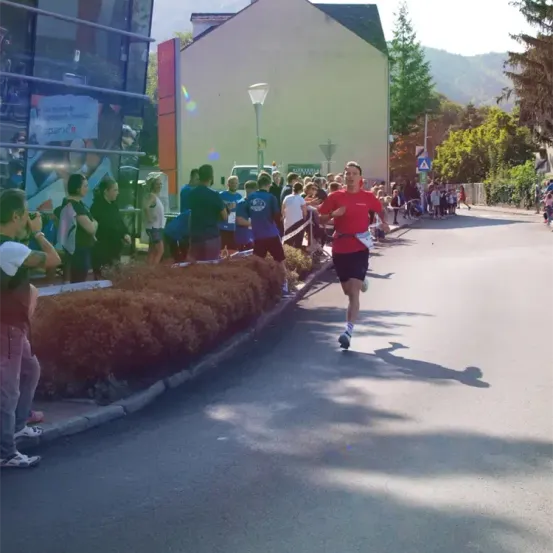Ein Mann läuft in einem Marathon, während mehrere Leute von der Straßenseite aus zusehen. Es gibt Gebäude und Bäume am Straßenrand.