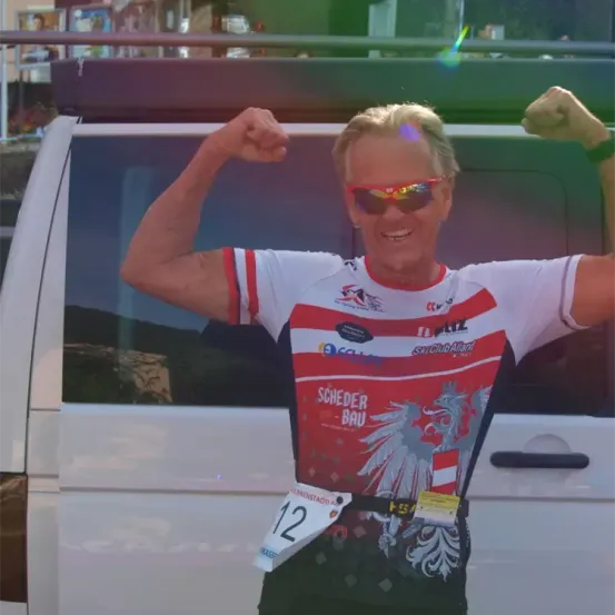 Ein Mann mit der Startnummer 12 steht neben einem Auto und zeigt seine Muskeln. Er trägt eine Sonnenbrille, eine Uhr und ein rot-weißes Trikot.