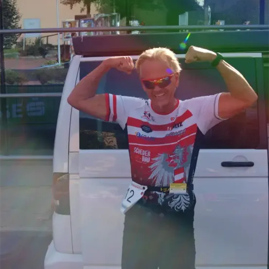 Eine Frau mit einem roten und weißen Radtrikot streckt ihre Muskeln nach außen. Sie steht vor einem weißen Van und hinter ihr befindet sich ein Baum und ein Schild.