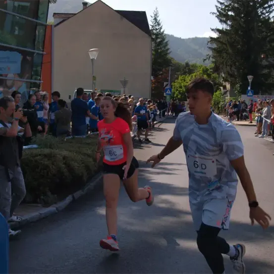 Eine junge Frau und ein junger Mann laufen einen Marathon auf einer Straße. Zuschauer säumen die Straße und feuern sie an. Ein Schild mit der Nummer 6D ist auf dem Shirt des Mannes sichtbar.