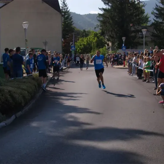 Ein Marathonläufer in Blau sprintet die Straße hinunter, umgeben von einer Menge von Zuschauern, mit Gebäuden und Bäumen im Hintergrund.