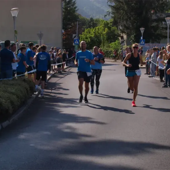 Ein Marathon findet statt, mit Läufern, die an Zuschauern auf der Seite vorbeilaufen. Die Läufer tragen nummerierte Startnummern. Bäume und Straßenlaternen sind im Hintergrund.