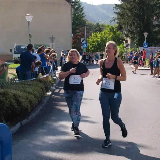 Zwei Frauen laufen in einem Marathon. Eine trägt die Nummer 7C und die andere 7D. Hinter ihnen stehen Menschen und beobachten.