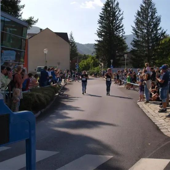Zwei Läufer überqueren die Ziellinie eines Marathons, mit Zuschauern auf beiden Seiten der Straße. Im Hintergrund sind Bäume und Häuser zu sehen.