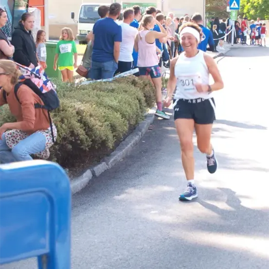 Eine Frau mit der Nummer 301 auf ihrem Shirt läuft einen Marathon, während Zuschauer sie anfeuern. Im Hintergrund steht ein grüner Mülleimer.