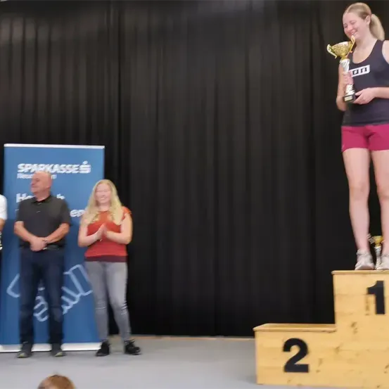 Eine Frau steht auf einem Podest und hält eine Trophäe in der Hand, mit einem Banner im Hintergrund. Hinter ihr stehen zwei Personen, eine klatscht und die andere beobachtet.