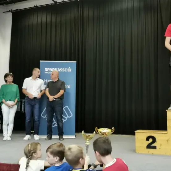 Drei Männer stehen auf einem Podest mit zwei Trophäen. Eine Frau steht neben ihnen und mehrere Kinder sitzen vorne. Ein Banner mit Logo steht dahinter.