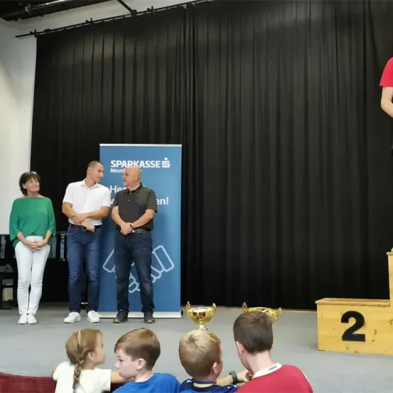 Drei Erwachsene und mehrere Kinder stehen mit Trophäen auf der Bühne. Ein Banner hinter ihnen trägt den Text 'Sparkasse'. Eine Person steht auf einem Podest rechts.