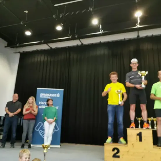 Eine Gruppe von Menschen steht mit Trophäen auf einer Bühne. Drei Männer stehen auf dem Podium, einer hält eine Trophäe. Zwei Frauen und ein Mann stehen hinter ihnen. Einer der Männer klatscht.