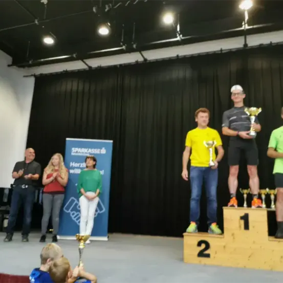 Drei Männer stehen auf einem Podest und halten Trophäen in der Hand, mit zwei Frauen, die hinter einem Banner klatschen. Ein Mann trägt ein gelbes T-Shirt, ein anderer ein graues T-Shirt und der dritte ein grünes T-Shirt. Eine Gruppe von Menschen steht auf der Bühne, darunter ein Mann in einem schwarzen T-Shirt.