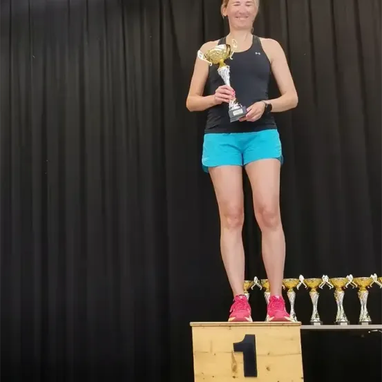 Eine Frau steht auf einem Holzpodium mit einer Trophäe, lächelt und trägt ein schwarzes Tanktop, blaue Shorts und pinke Schuhe.