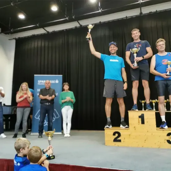 Drei Männer stehen auf einem Podest und halten Trophäen. Der Mann auf der linken Seite hält seine Trophäe hoch. Zuschauer stehen hinter dem Podest und klatschen. Ein Banner zeigt 'SPAR' und 'SSE'.