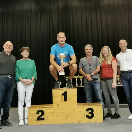 Ein Mann auf einem Podium mit einer Trophäe steht mit drei anderen Männern und zwei Frauen, alle lächeln, vor einem schwarzen Vorhang.