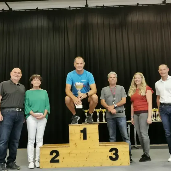 Sechs Personen stehen auf einem Podest, einer Mann auf dem ersten Platz, hält einen Pokal. Sie sind lässig gekleidet, lächeln und stehen auf einer Bühne mit schwarzem Vorhang als Hintergrund.