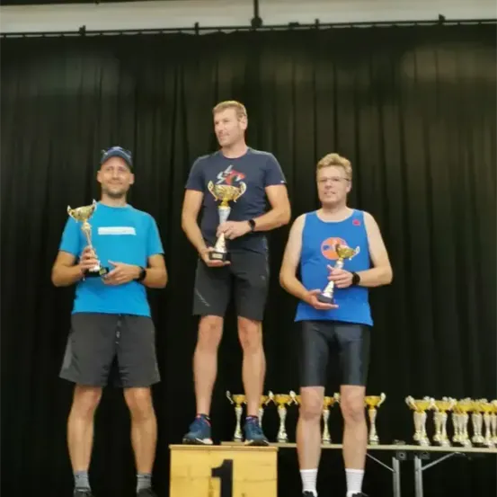 Drei Männer stehen auf einem Podest und halten Pokale. Einer trägt ein blaues T-Shirt, ein anderer ein schwarzes T-Shirt und der dritte ein blaues Tanktop. Sie lächeln und schauen in die Kamera.