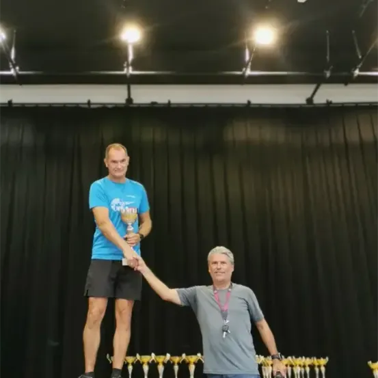 Ein Mann steht auf einem Podium und hält einen Pokal, während ein anderer Mann seine Hand hält. Hinter ihnen ist ein schwarzer Vorhang mit zwei Lichtern an der Decke.