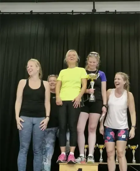 Eine Gruppe von Frauen steht auf einer Bühne, zwei von ihnen halten Pokale. Sie lächeln und scheinen zu feiern. Sie stehen auf einem Podest mit der Nummer 1. Dahinter ist ein schwarzer Vorhang und Pokale auf einem Tisch.