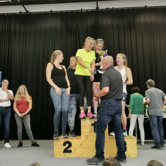 Eine Gruppe von Frauen in Turnschuhen und Freizeitkleidung steht auf einem Podest mit einer Trophäe. Das Podest hat die Nummer 2 darauf. Ein Mann steht vor ihnen. Hinter ihnen ist ein schwarzer Vorhang und einige Leute stehen vor einem Tisch.