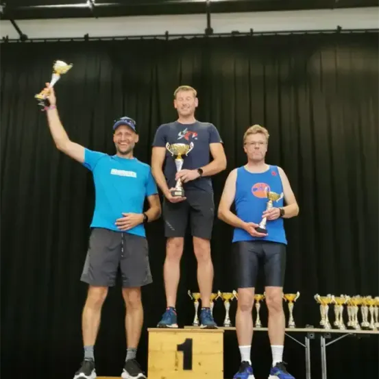 Drei Männer stehen auf einem Podest mit Trophäen. Sie lächeln und scheinen zu feiern. Sie stehen auf einer Bühne mit schwarzem Hintergrund.