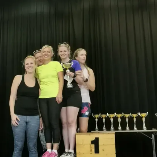 Fünf Frauen stehen auf einem Podest mit der Nummer 1 und lächeln. Sie halten einen Pokal. Dahinter stehen Pokale und ein schwarzer Vorhang.