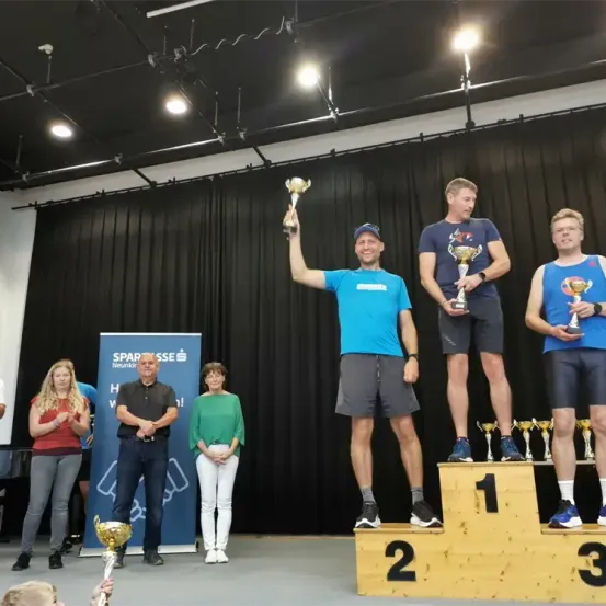 Drei Männer stehen auf einem Podium, der in der Mitte hält eine Trophäe und lächelt. Zwei andere Männer halten Trophäen in den Händen. Eine Frau und ein Mann stehen hinter ihnen, und mehrere Trophäen sind auf dem Podium. Ein Banner mit dem Text 'SPARÖSSE Neunkir' ist auf der linken Seite.