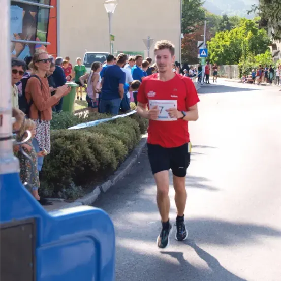 Ein Mann in einem roten Shirt und schwarzen Shorts läuft einen Marathon. Zuschauer stehen am Straßenrand, einige halten Schilder. Bäume und Gebäude sind im Hintergrund.