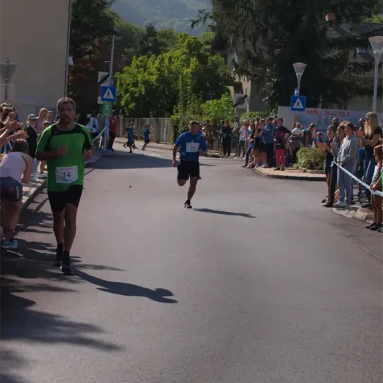 Läufer nehmen an einem Marathon teil, wobei Nummer 14 in Führung liegt. Zuschauer säumen die Straße auf beiden Seiten und feuern an. Bäume und Straßenlaternen sind im Hintergrund zu sehen.