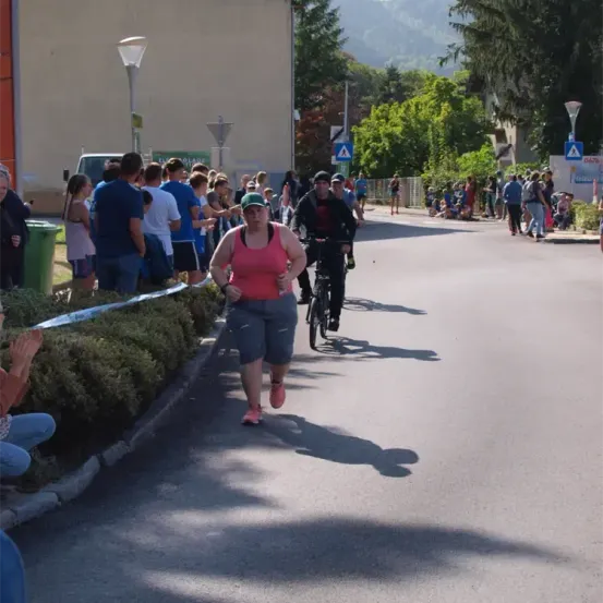 Eine Gruppe von Menschen steht an einer Straße und beobachtet eine Frau in einem rosa Tanktop und einen Mann auf einem Fahrrad. Einige Leute sitzen und stehen auf dem Bürgersteig.