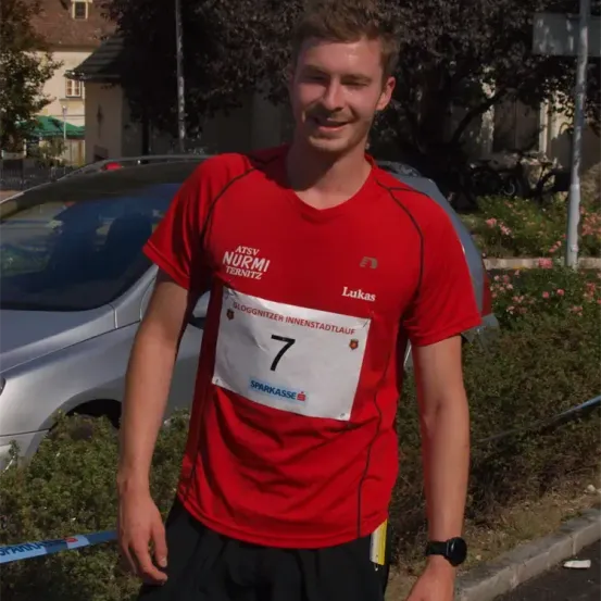 Ein Mann steht vor einem Auto und trägt ein rotes T-Shirt mit weißen Nummern auf der Vorderseite. Er lächelt in die Kamera.