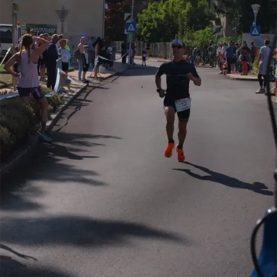 Ein Mann in einem schwarzen und orangefarbenen Outfit läuft bei einem Marathon mit der Nummer 12 auf dem Rücken. Zuschauer beobachten von der Seite.