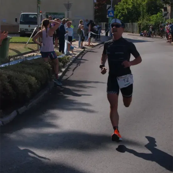 Ein Mann in einem Laufoutfit mit der Nummer 12 läuft auf einer Straße mit Zuschauern und einem Fahrzeug im Hintergrund.