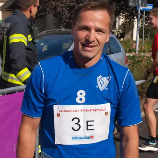 Ein Mann in einem blauen Sporttrikot mit der Nummer 8 lächelt in die Kamera. Er hat die Startnummer 3E auf seinem Trikot. Hinter ihm steht ein Feuerwehrmann in Uniform.