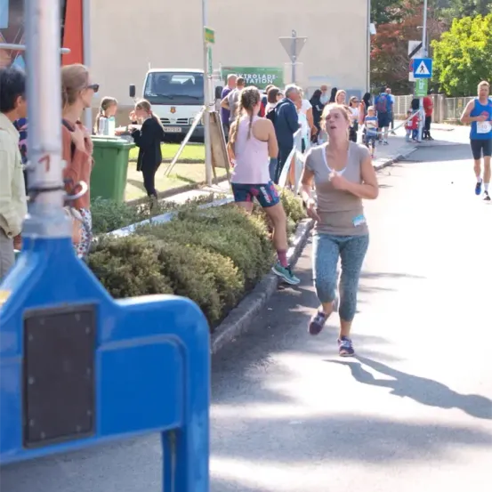 Teilnehmer eines Rennens sind auf einer Straße zu sehen, während andere zuschauen. Eine Frau läuft mit offenem Mund, und ein Mann läuft vor ihr. Eine Frau im Hintergrund schaut die Läuferin an. Es gibt einen grünen Mülleimer und ein Gebäude im Hintergrund.