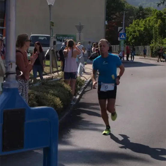 Ein Mann läuft bei einem Marathon. Er trägt ein blaues Shirt und grüne Schuhe. Zuschauer stehen am Straßenrand. Bäume und ein Gebäude im Hintergrund.