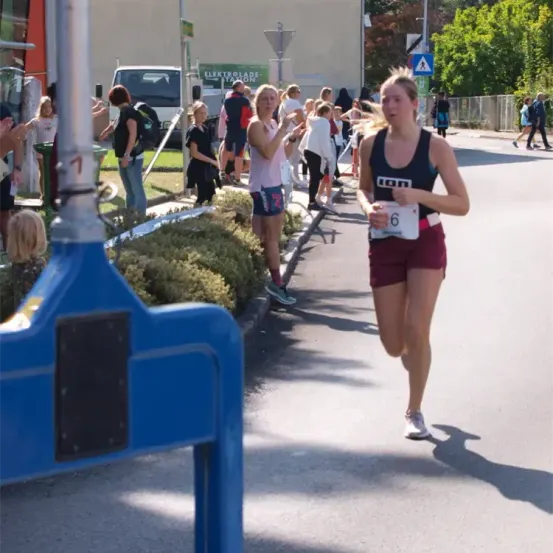 Eine Läuferin mit der Nummer 6 läuft auf einer Straße, die von Zuschauern beobachtet wird. Sie trägt ein Tanktop und Shorts. Hinter ihr steht ein blauer Pfosten.