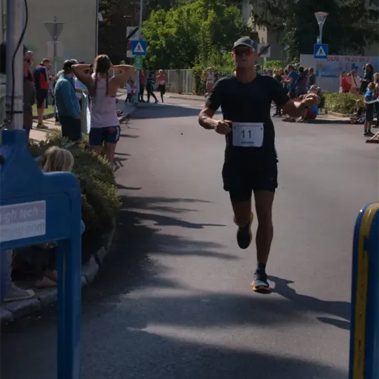 Ein Mann mit der Nummer 11 auf dem Trikot läuft einen Marathon. Zuschauer säumen die Straße und feuern ihn an. Im Hintergrund sind Bäume und Straßenlaternen zu sehen.