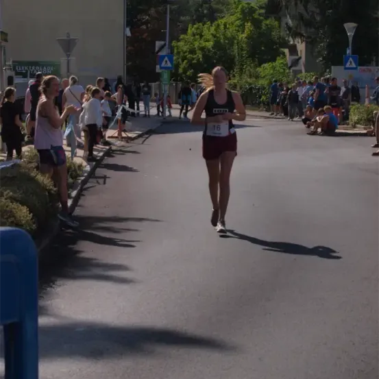 Eine Läuferin mit der Nummer 16 auf ihrem Tanktop läuft bei einem Marathon. Zuschauer säumen die Straße, einige sitzen auf dem Bürgersteig, andere stehen. Der Schatten der Läuferin wird auf die Straße geworfen.