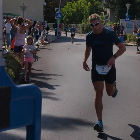 Ein Mann läuft in einem Marathon, Nummer 3 auf seinem Trikot, während Zuschauer von der Straßenseite zuschauen. Ein Mädchen und eine Frau in der Nähe beobachten ebenfalls.