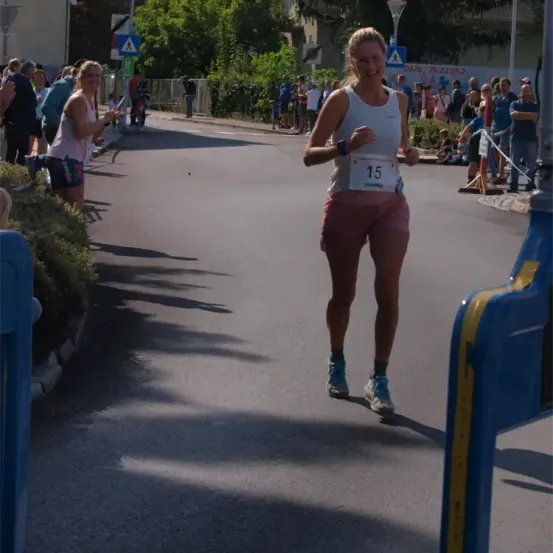 Eine Läuferin mit der Nummer 15 läuft eine Straße hinunter, während Zuschauer zusehen. Es gibt Büsche und ein Gebäude an der Seite.